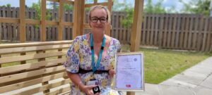 Shirley Cochrane sat on a bench, in a garden, with her certificate and pin badge