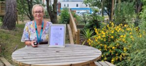 Shirley Cochrane sat at a table with Colchester Hospital in the background, with her certificate and pin badge