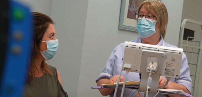 A patient wearing a mask, talks to a nurse who is also wearing a mask.
