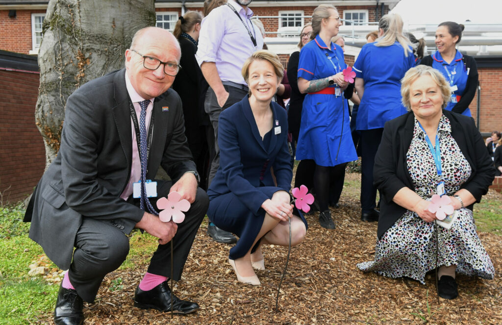 Amanda Pritchard (centre) with ESNEFT Chief Executive Nick Hulme (left) and ESNEFT Chair Helen Taylor, all crouching, holding a pink, iron imitation blossom.